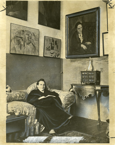 Gertrude Stein sitting on a sofa in her Paris studio. Notice her portrait painted by Picasso in the upper right corner. Photo by World Wide Photos (May 1930). PD-100+. Wikimedia Commons. 