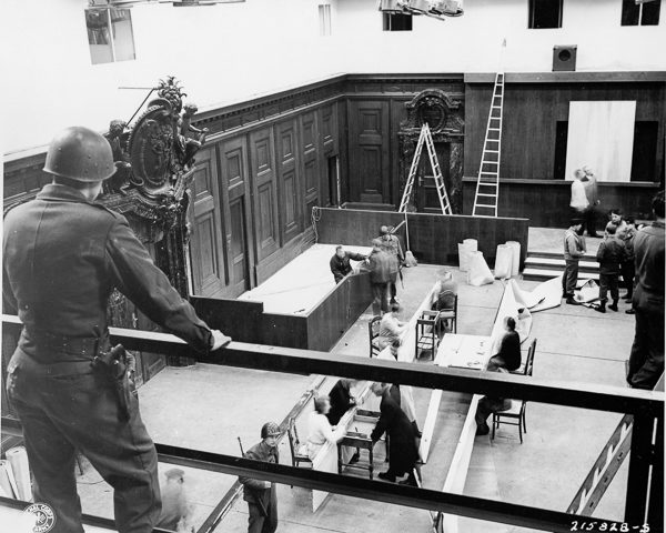 PFC Carl D. Sander oversees repairs to the courtroom prior to the Nuremberg Trials. The defendants’ dock is visible in the center of the photo. The windows in the top of the photo were specially installed for photographers and movies. Photo by US Army (c. 1945). PD-US Government. Wikimedia Commons.