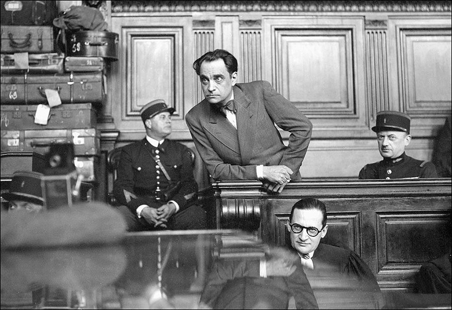 Marcel Petiot (upright, center) stands trial in Paris in March 1946. Notice the suitcases to the left of Petiot and the policeman. Photo by anonymous (c. March 1946).