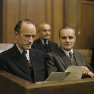 Alfried Krupp (left) reading a document, seated in the dock, as a defendant in the Krupp Trial. Photo by anonymous (c. 1948). Courtesy of Stadtarchiv Nürnberg. Wikimedia Commons. 