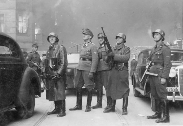 The leader of the grand operation (original title). Jürgen Stroop (center) and Josef Blösche (far right) standing watching the burning of a Warsaw townhouse (Nowolipie 62) next to the ghetto wall. Photo by Franz Konrad or Propaganda Kompanie nr 689 (c. April/May 1943). The Stroop Report. U.S. National Archives and Records Administration and Polish Institute of National Remembrance. PD-Expired Copyright. Wikimedia Commons.
