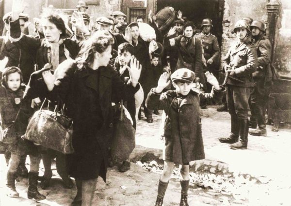 A Jewish boy surrenders in Warsaw (original title). Polish Jews are captured by Germans during the suppression of the Warsaw Ghetto Uprising. Josef Blösche stands (right with goggles on helmet) with machine gun pointed at the young boy. Photo by Franz Konrad or Propaganda Kompanie nr 689 (c. April/May 1943). The Stroop Report. U.S. National Archives and Records Administration and Polish Institute of National Remembrance. PD-Expired Copyright. Wikimedia Commons.