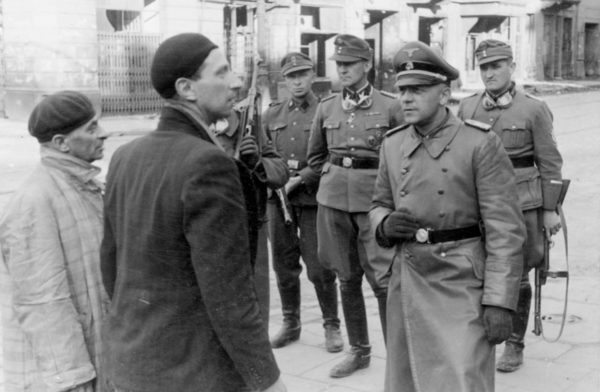 Jewish Traitors (original title). SS officer questions two Jewish resistance fighters as Jürgen Stroop (rear center) listens. Photo by Franz Konrad or Propaganda Kompanie nr 689 (c. April/May 1943). The Stroop Report. U.S. National Archives and Records Administration and Polish Institute of National Remembrance. PD-Expired Copyright. Wikimedia Commons.