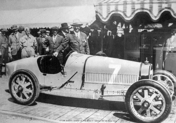 Ettore Bugatti (center) standing next to (behind) a Bugatti Type 35, the same car which W. Williams raced. This car participated in the Grand Prix de Lyon. Photo by anonymous (c. 1924). Photo posted by Arnaud 25. PD-CCA-Share Alike 4.0 International. Wikimedia Commons.