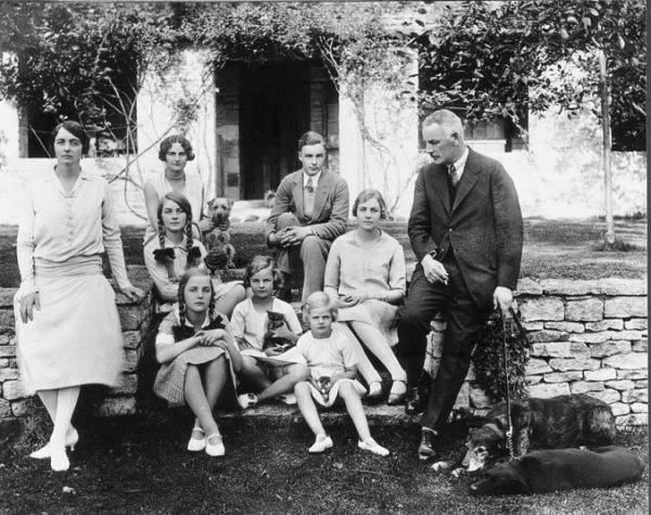 The Mitford family. Sydney Bowles (far left) married to David Freeman-Mitford (far right). Unity Mitford is sitting in front row on the step (left) while Jessica Mitford is sitting next to her on the right holding a cat. Diana Mitford is sitting in second row behind Unity. Photo by anonymous (c. 1928). PD-UK artistic work of which author is unknown. Wikimedia Commons.