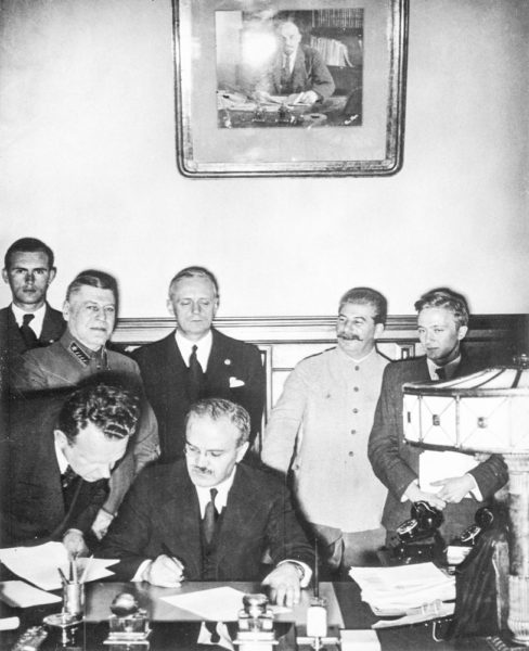 Soviet Foreign Minister V. Molotov signs the German-Soviet Treaty of Friendship (Molotov-Ribbentrop Pact). Behind Molotov stands Joachim von Ribbentrop. To the right of Ribbentrop is Joseph Stalin. Photo by anonymous (23 August 1939). National Archives & Records Administration. PD-Russian Public Domain. Wikimedia Commons.