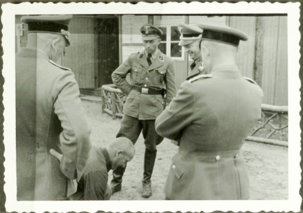 A KZ Sachsenhausen inmate kneels in front of four SS henchmen, including Commander Karl Otto Koch (second from right) who laughs at him. Photo by anonymous (c. 1937). PD-Author Life + 70. Wikimedia Commons.