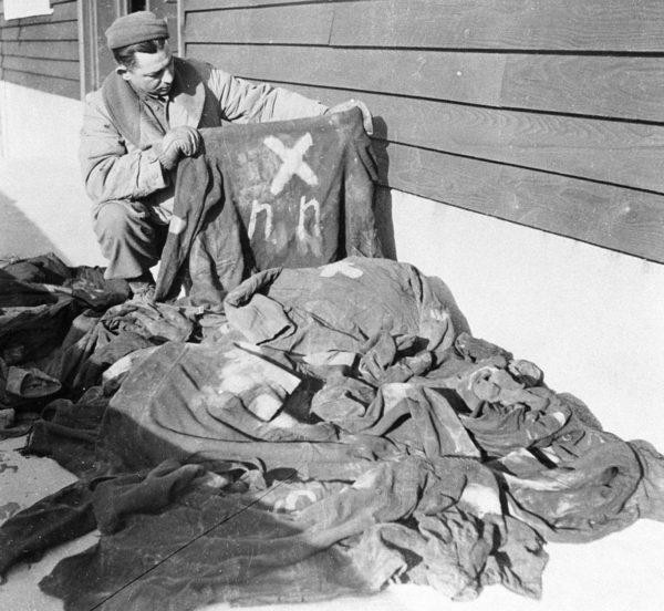 A United States soldier looks at prisoner clothing that is marked NN for “Nacht und Nebel.” Photo by anonymous (c. December 1944). United States Holocaust Memorial Museum. PD-U.S. Government. Wikimedia Commons. 