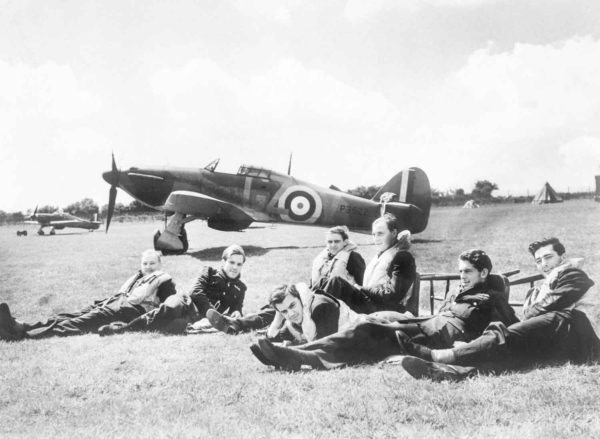 Pilots of “B” Flight, No. 32 Squadron relax on the grass at Hawkinge in front of a Hurricane fighter plane. All survived the Battle of Britain except for Pilot Officer Keith Gillman (second from left). Photo by anonymous (29 July 1940). Imperial War Museum. PD-Expired copyright. Wikimedia Commons. 