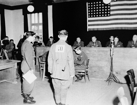 Former SS-Hauptsturmführer Friedrich Wetzel (1909−?) standing in front of the judges at the Dachau/Buchenwald trials. Wetzel was convicted and is listening to his death sentence being pronounced. He was released in 1952. Photo by anonymous (13 December 1945). PD-U.S. Government. Wikimedia Commons. 