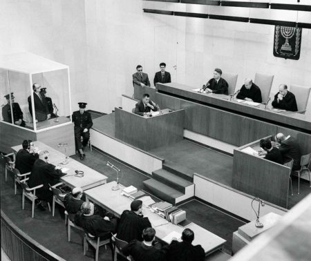 Defendant Adolf Eichmann (standing inside glass booth) is sentenced to death. Photo by anonymous (15 December 1961). National Photo Collection of Israel. PD-Expired copyright. Wikimedia Commons.