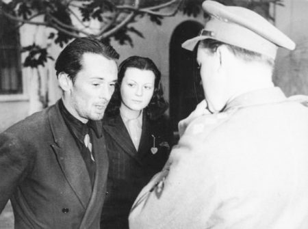 John Amery (left) surrendering to British officials. His mistress, Michelle Thomas (center), looks on. To the right, facing away from the camera, is Capt. Alan Whicker. Photo by anonymous (c. 1945). Bruce Quarry collection. PD-Italian Public Domain. Wikimedia Commons.