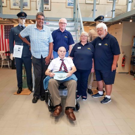 Stan Booker (seated) and members of the FONFA Heritage Museum. Standing from left to right: Sam Ruddy (archivist), Dave Connor (volunteer), Margaret Williams (members’ secretary), and Steve Williams (trustee). To the left of Sam Ruddy is Stan’s RAF uniform that he donated to the museum. Photo by Pat Vinycomb (c. October 2023). Courtesy of Pat Vinycomb.