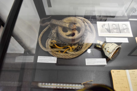 Artifacts from the Petiot criminal case. Upper left: the pulley and rope with iron chain and hook used by Petiot to lower his victims into the pit. Upper right: Petiot’s mug shots. The viewing lens can be seen next to the rope and below the mug shots. Photo by Sandy Ross (c. September 2022). Paris Police Museum.